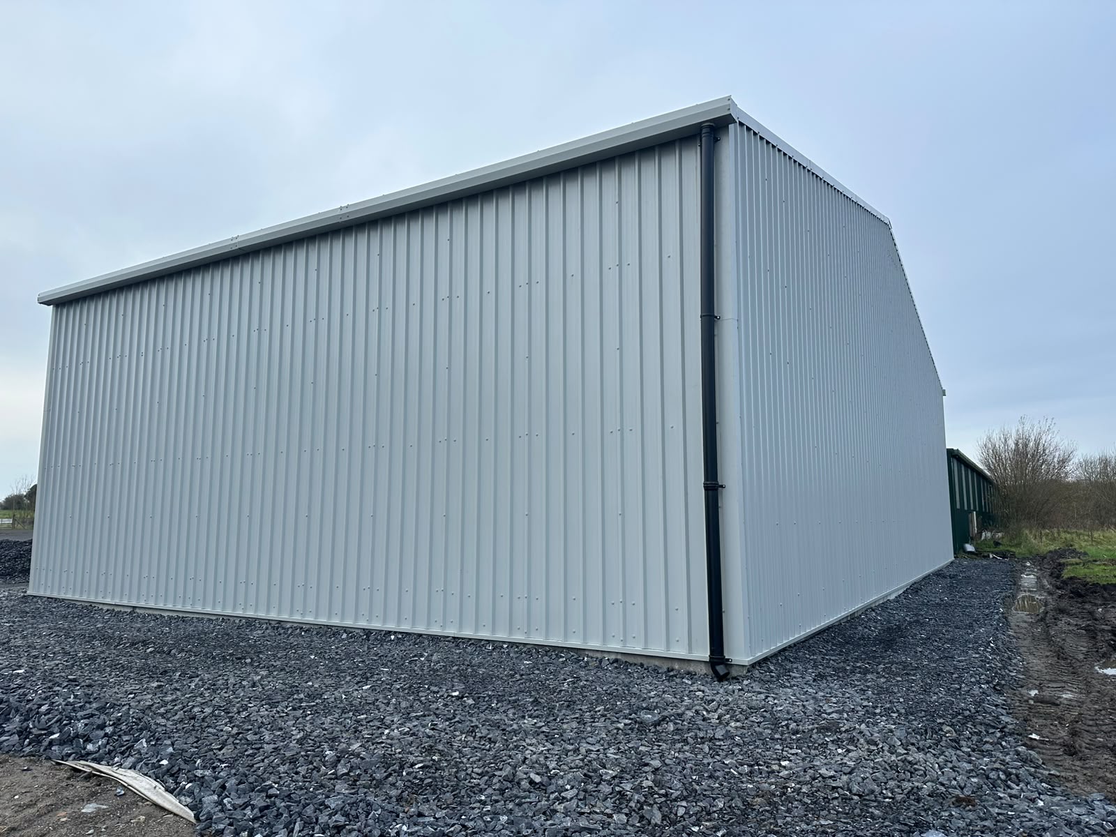 Completed pale grey clad steel shed on new stone hardcore, showing gable end and downpipe