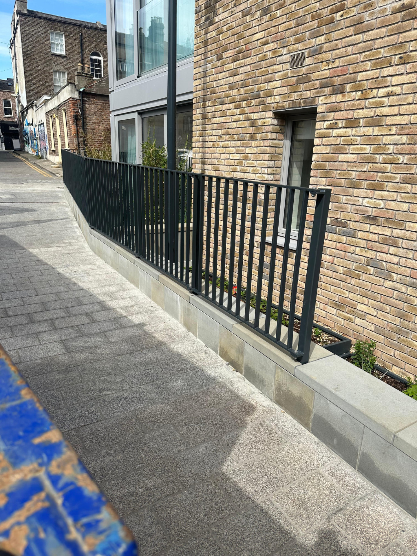 Run of dark grey vertical bar boundary railings on a low stone plinth along a footpath beside a brick apartment building