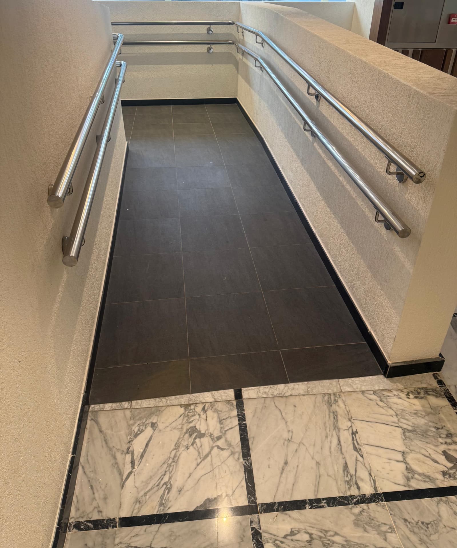 Polished stainless steel handrails to both sides of an internal access ramp in a tiled hotel lobby