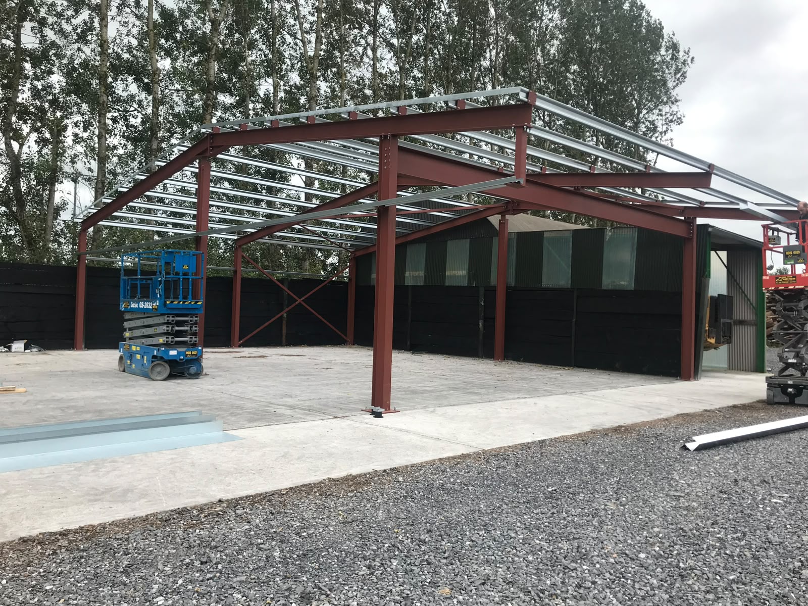 Red oxide steel portal frame with galvanised purlins erected over a concrete yard beside an existing shed