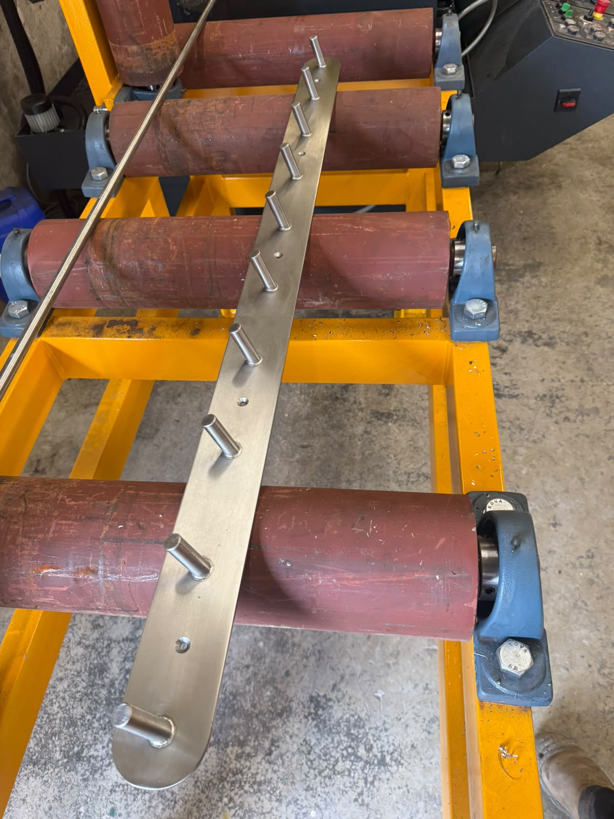 Stainless steel bar with welded and dressed pins set out along it, resting on workshop rollers