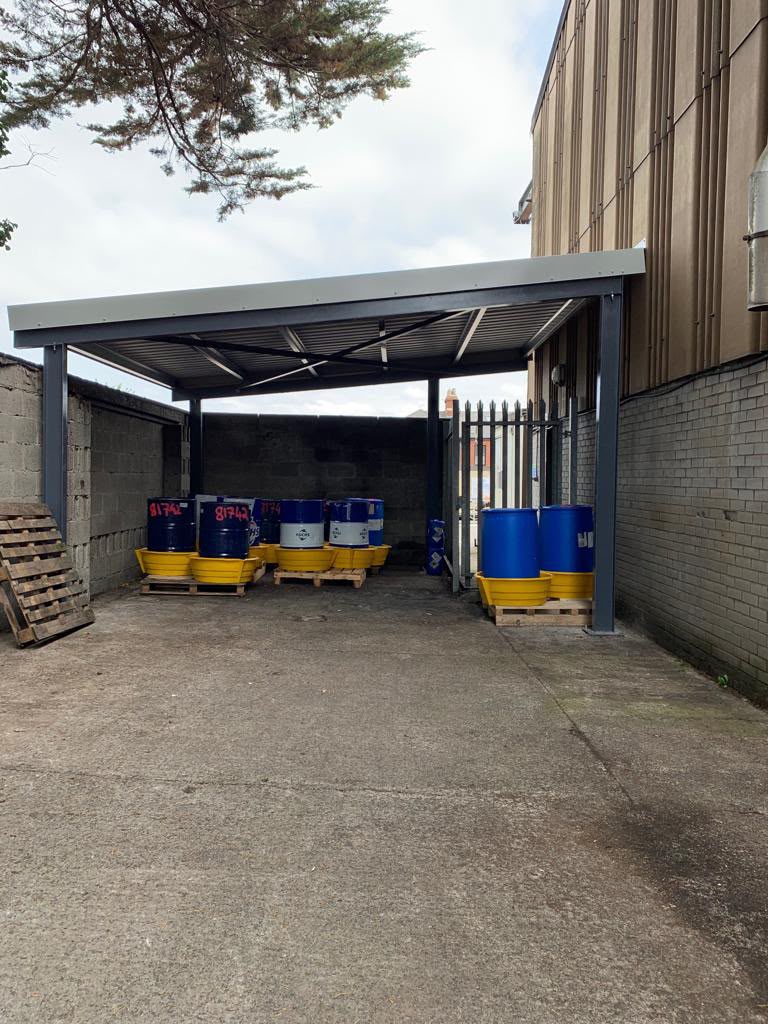 Dark grey steel canopy with bracing and translucent roof sheeting over a bunded drum storage area