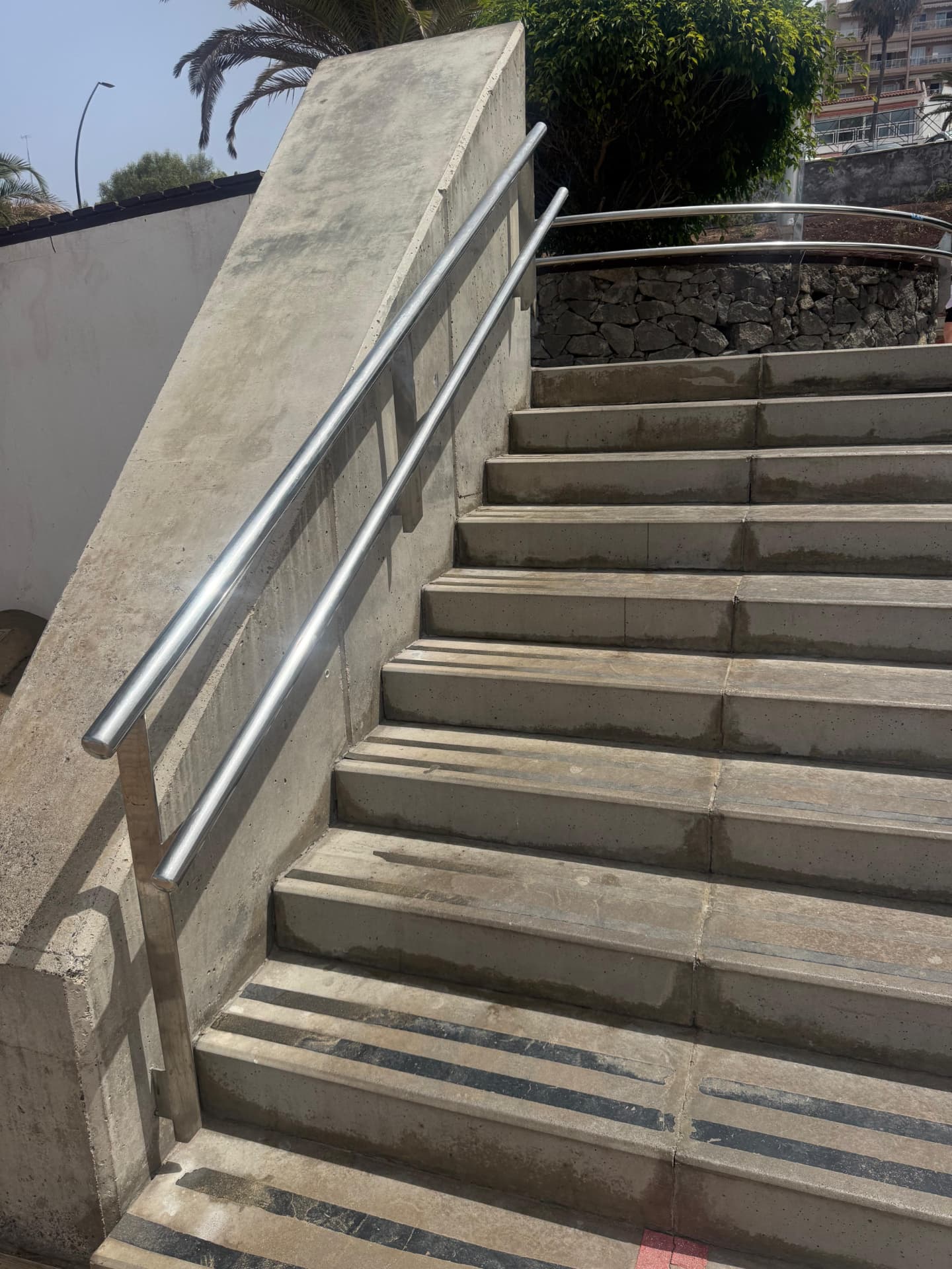Two run stainless steel handrail fixed to a concrete wall alongside a wide external concrete staircase