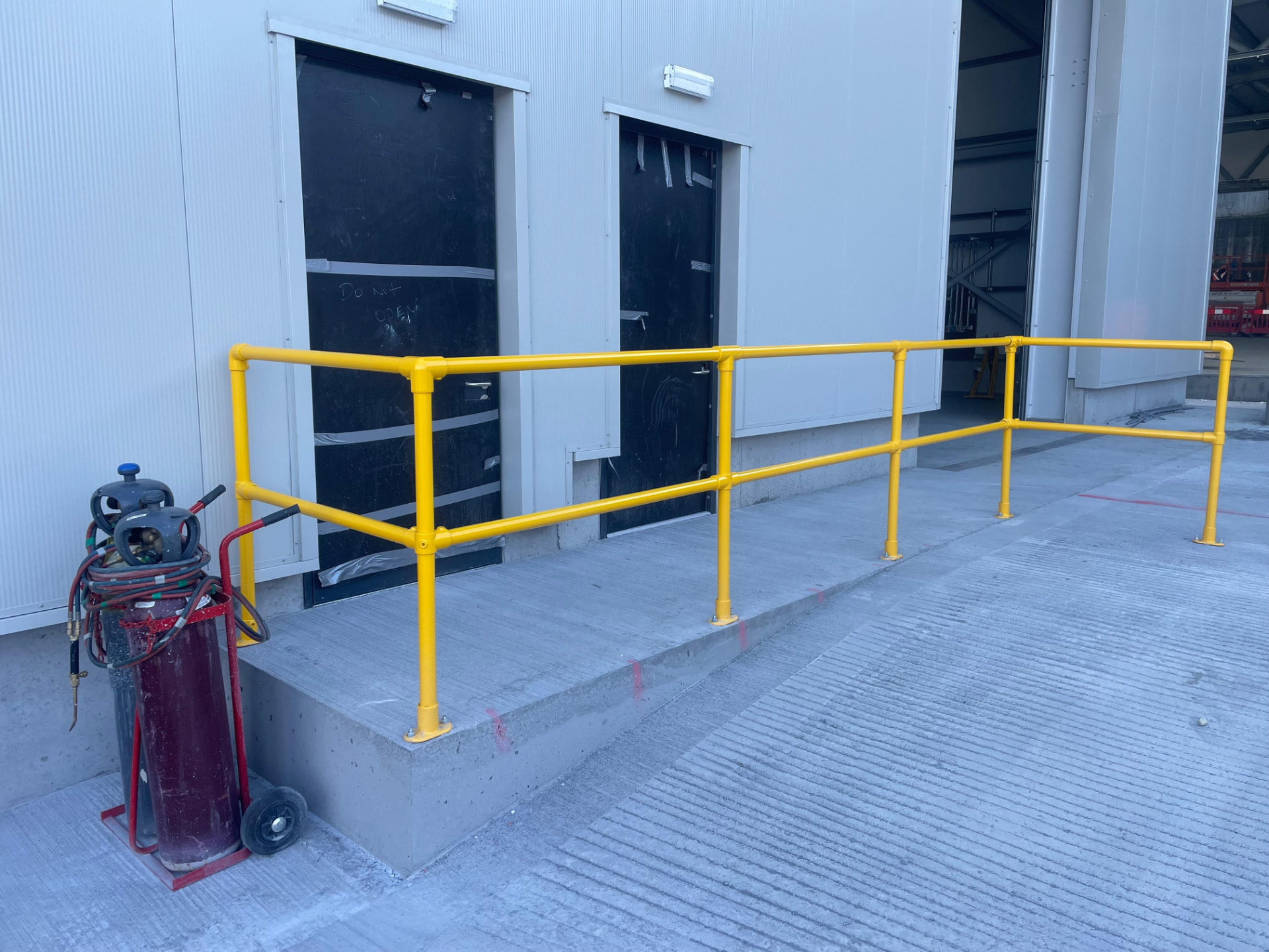 Yellow handrail with base plated posts protecting a raised concrete plinth outside personnel doors