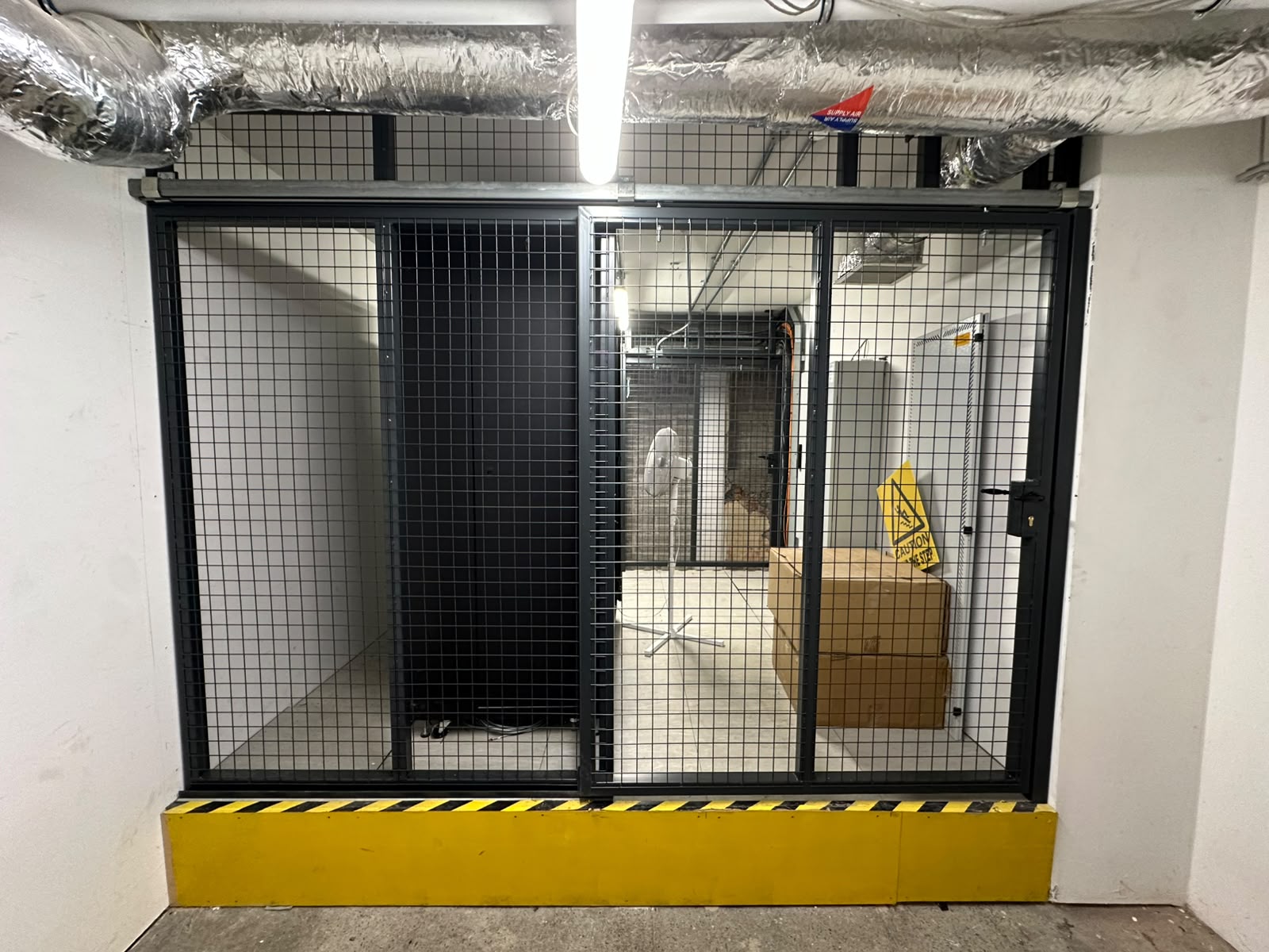 Dark grey welded mesh cage with sliding doors and lock securing a basement plant room, with yellow kerb protection